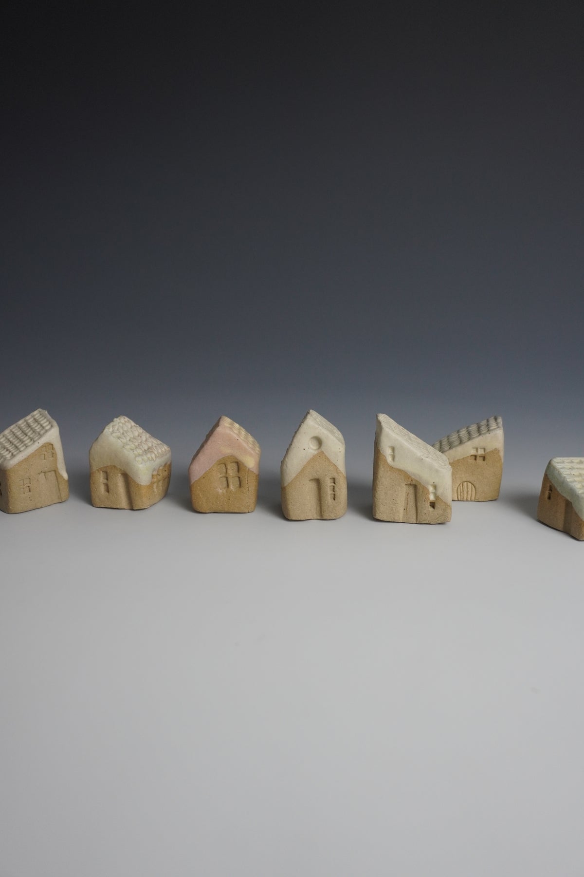 A row of small hand-carved wooden house figurines in natural beige wood, arranged on a light gray surface against a dark gray gradient background.