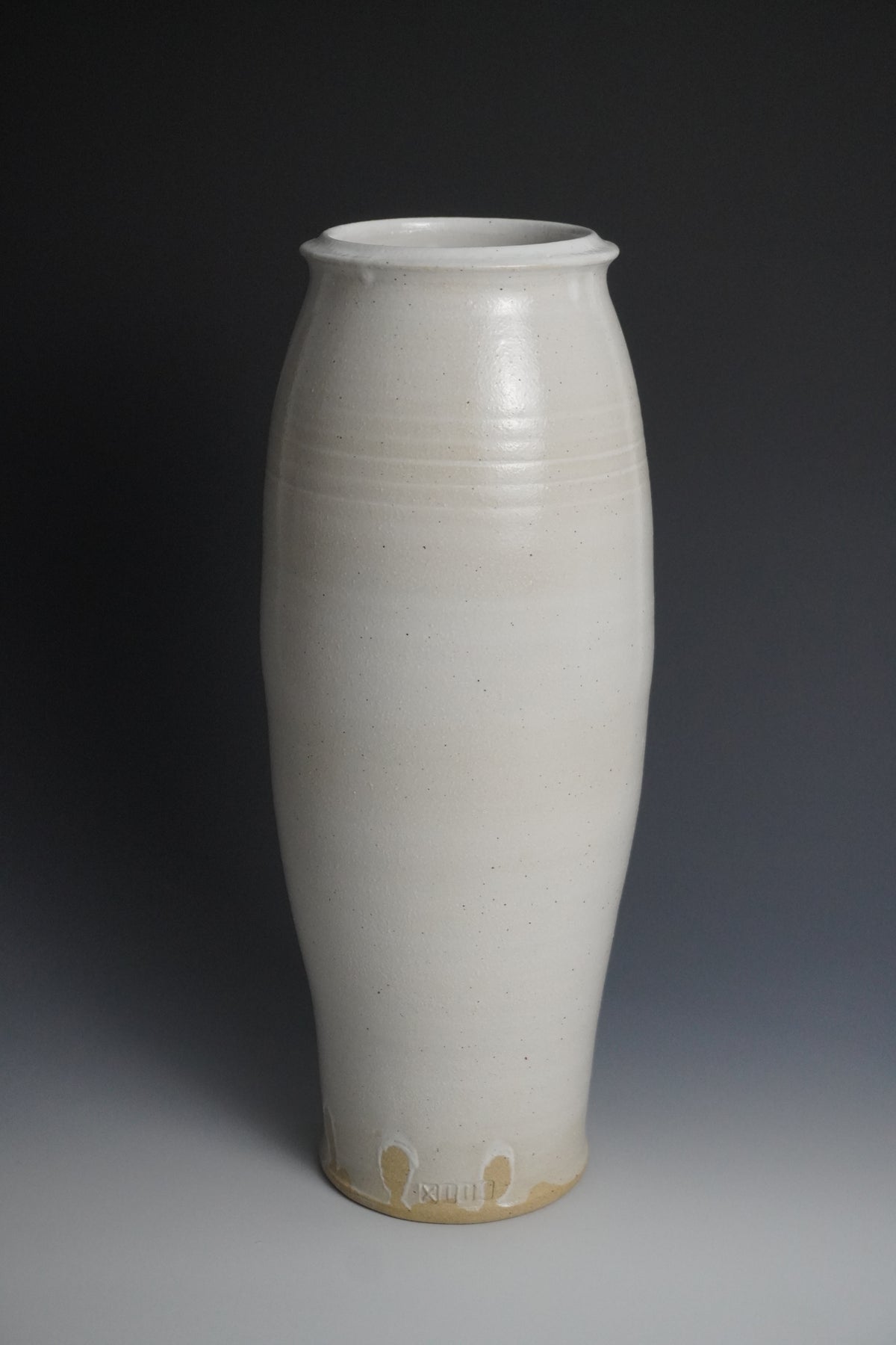 A tall, slender ceramic vase with a matte off-white glaze and a natural beige band at the base, photographed against a dark gray studio background.