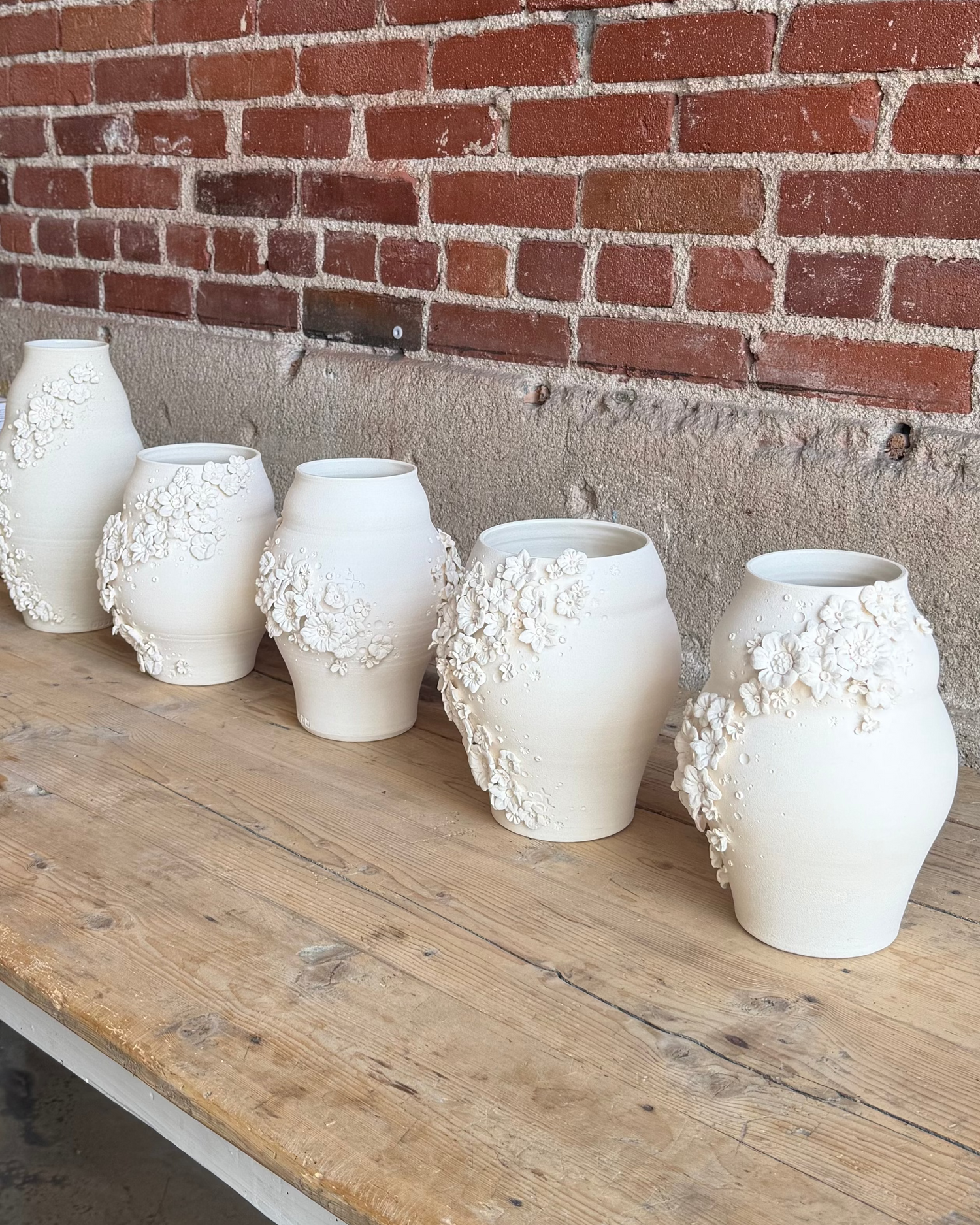 Row of white ceramic vases on a wooden table against a brick wall.