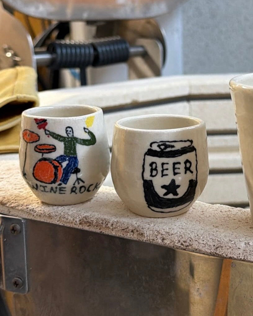 two hand painted shot glasses. decorated with glaze and shiny. the decoration is of a beer can and the other is a funny drawing of a person playing the drums that says "wine rocks"