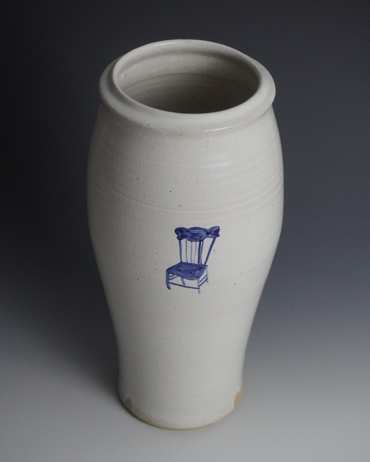 A white stoneware vase with a blue printed chair design on the front, photographed against a dark-to-light gray studio background.