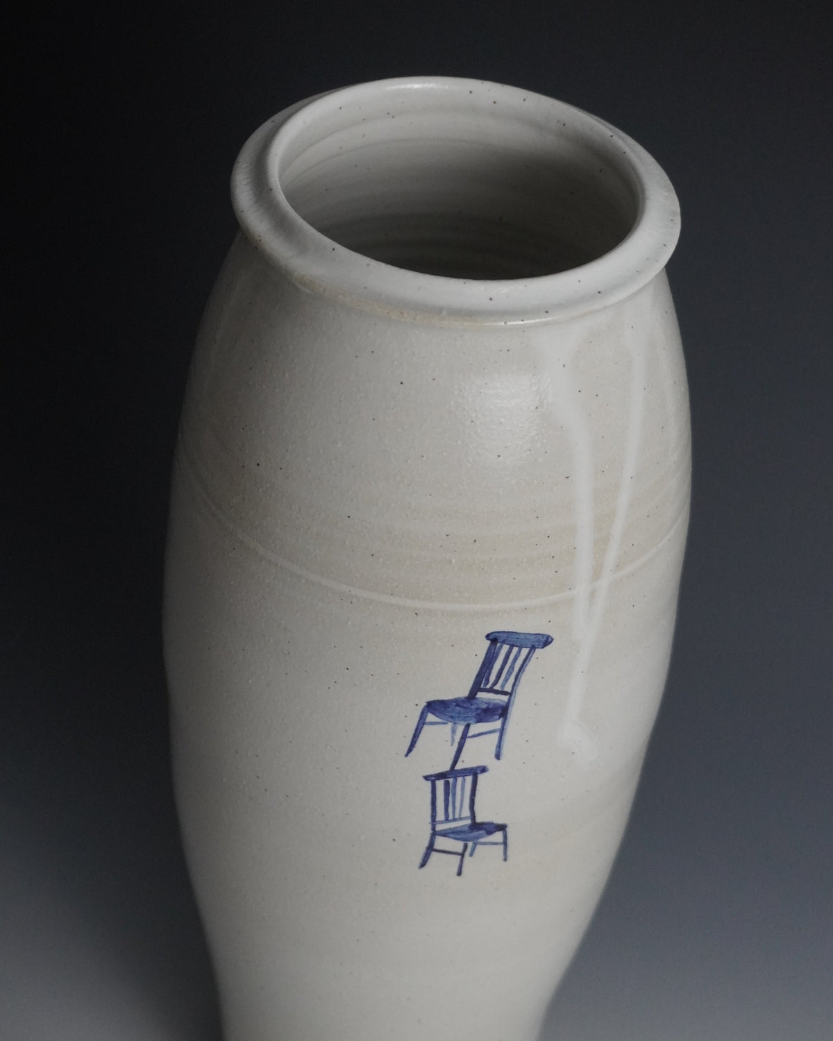 A handmade ceramic vase with a matte off-white glaze and a blue painted illustration of two stacked chairs, set against a dark gray gradient background.