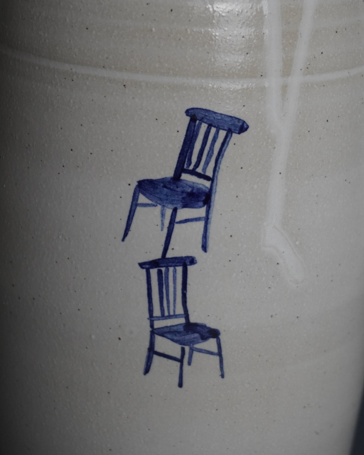 Close-up of a speckled off-white ceramic surface featuring a hand-painted blue illustration of two stacked chairs.