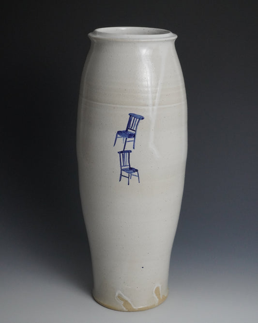 A tall, slender ceramic vase with a matte off-white glaze and a blue painted illustration of two stacked chairs on the front, set against a dark gray studio background.